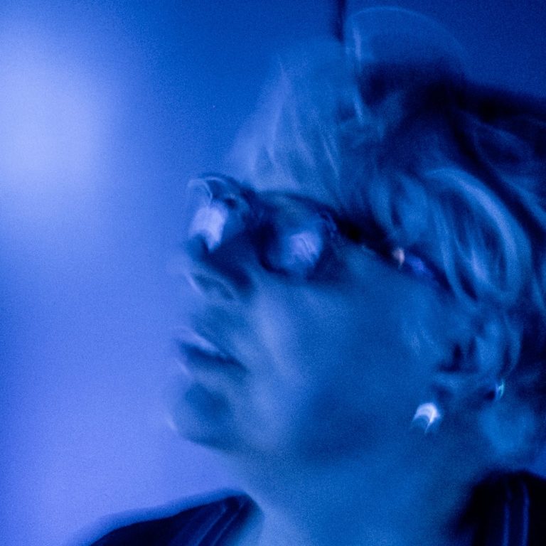 Eine porträtartige Darstellung einer Person mit Brille, in blauer Beleuchtung.